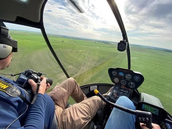 View from helicopter cockpit over green fields, with two people inside, one holding a camera.