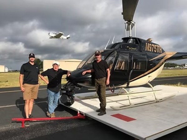 Three men stand by a helicopter on an airstrip, with a small plane flying in the background.