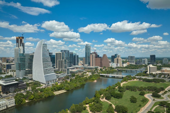 City skyline with modern buildings, river, and park under a blue sky with clouds.